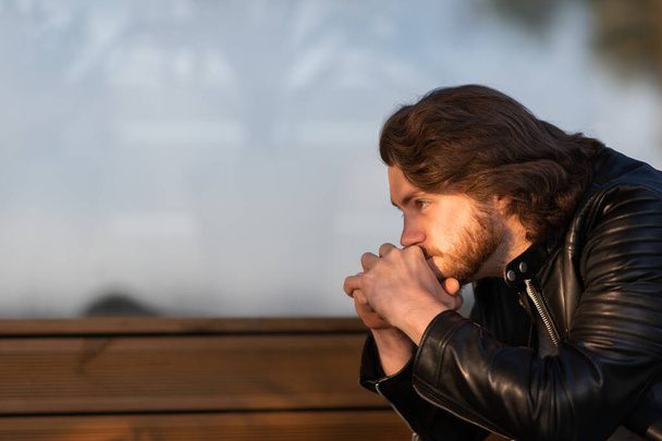 Серйозний молодий бородатий чоловік у чорній шкіряній куртці сидить і думає на відкритому повітрі
 - Фото, зображення