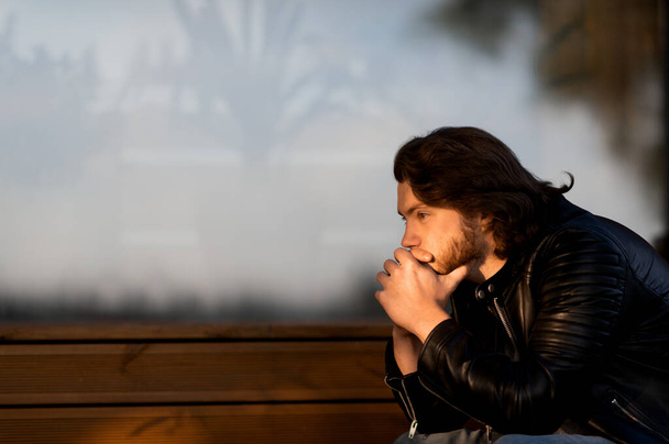 Pensive poważne młody brodaty mężczyzna nosić czarny skórzana kurtka siedzi i myśli na zewnątrz - Zdjęcie, obraz