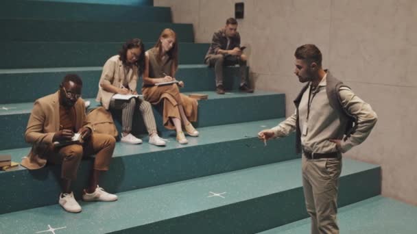 Отслеживание медленного снимка современного профессора-мужчины, дающего урок многоэтничной группе студентов университета, сидящих на лестнице перед лекционным залом и делающих заметки в тетрадях - Кадры, видео