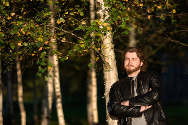Egy fiatal szakállas férfi áll a fák mellett az őszi parkban, és elgondolkodva néz a távolba. - Fotó, kép