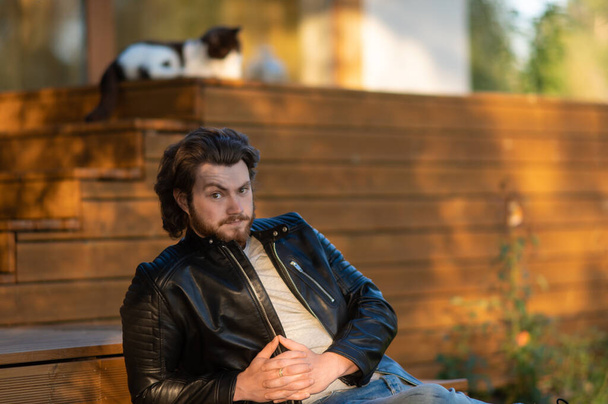 un joven barbudo con una chaqueta de cuero negro sentado en la terraza de la casa y mirando pensativamente a la cámara - Foto, imagen