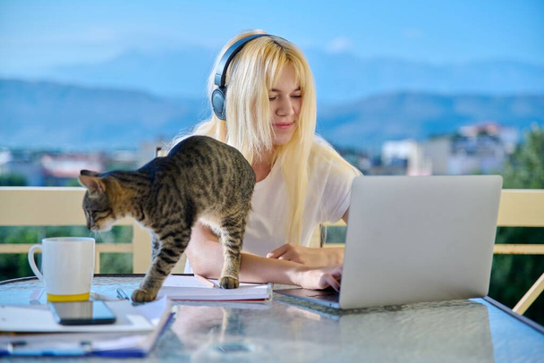 Female student studies at home online using laptop. Teenager sitting on outdoor balcony with pet cat in headphones, school notebooks, looking at screen. E-learning, modern technologies in education - Фото, изображение