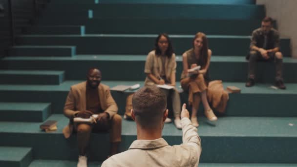 Медленная фотография профессора мужского пола, выступающего перед группой студентов многонациональных университетов, сидящих на бирюзовой лестнице в библиотеке или аудитории - Кадры, видео