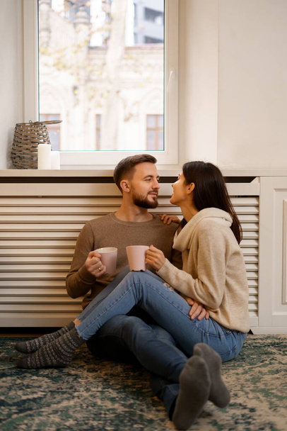 Romantic couple at home. An Attractive young woman and handsome man are enjoying spending time together while siting coddling with cups of tea in hands. - Photo, Image