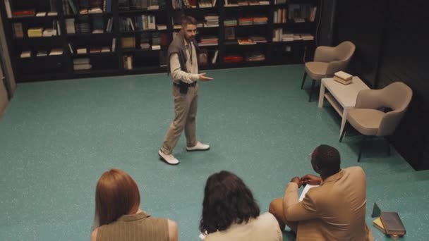 Z góry śledzenie powolne ujęcie współczesnego profesora uniwersytetu męskiego w inteligentnych ubraniach casualwear dając wykład do grupy studentów wieloetnicznych siedzi na schodach w nowoczesnej bibliotece - Materiał filmowy, wideo