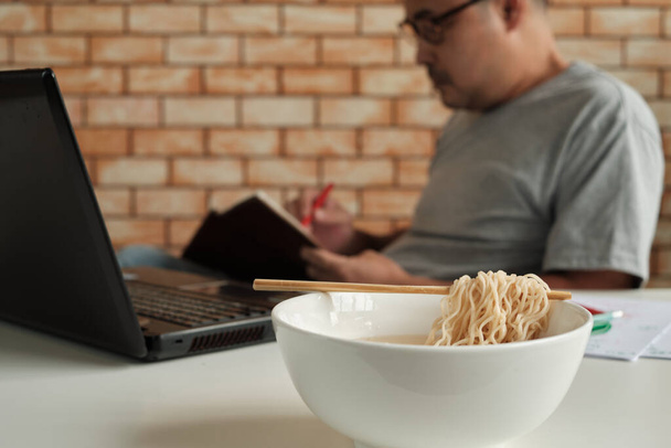 Тайский работник-мужчина занят работой с ноутбуком, используя палочки для еды поспешно есть лапшу во время перерыва на обед в офисе, потому что быстро, вкусно и дешево. Со временем азиатский фаст-фуд, нездоровый образ жизни. - Фото, изображение
