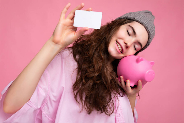 Okouzlující úsměv šťastný radostné mladé kudrnaté brunetky žena na sobě růžová košile a šedý klobouk izolované na růžovém pozadí s volným místem a držení růžové prase peníze box a kreditní karty pro mockup - Fotografie, Obrázek