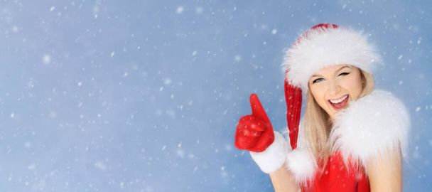 Young funny woman in a Santa Claus hat smiles winks and shows a like thumb with a red mitten - Photo, Image