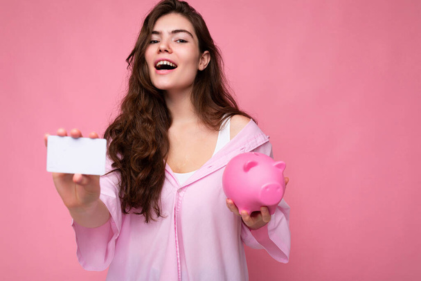 Kaunis onnellinen iloinen nuori brunette nainen yllään paita eristetty vaaleanpunainen tausta vapaata tilaa ja tilalla vaaleanpunainen sika rahaa laatikko ja luottokortti mockup - Valokuva, kuva