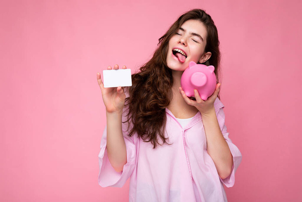 Atractiva joven morena feliz vistiendo camisa aislada sobre fondo rosa con espacio vacío y sosteniendo caja de dinero de cerdo rosa y tarjeta de crédito para maqueta - Foto, imagen
