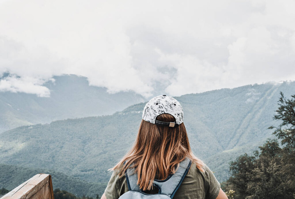 Visão traseira da jovem mulher loira em boné com mochila olhando para a vista das montanhas caucasianas e céu nublado em pé no ponto de vista de madeira, caminhadas estilo de vida saudável ativo apreciando a natureza - Foto, Imagem