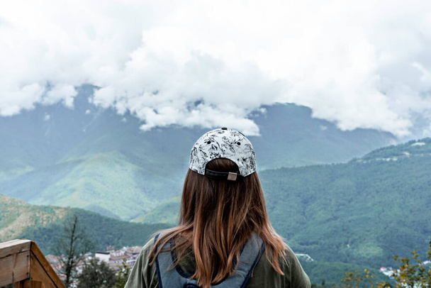 Вид сзади на молодую блондинку в кепке с рюкзаком, смотрящую на Кавказские горы и облачное небо, стоящее на деревянной смотровой площадке, гуляющую активный здоровый образ жизни, наслаждаясь природой - Фото, изображение
