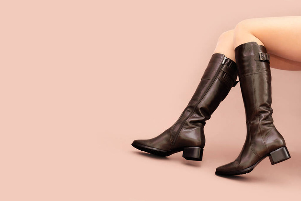 Caucasian female legs bent in knees in high brown boots on light dusty pink beige background. Copy space in left side, front view. - Fotó, kép