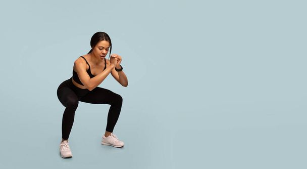 Trainingskonzept. Sportliche afrikanisch-amerikanische Dame macht tiefe Kniebeugen, Training vor blauem Hintergrund, Panorama - Foto, Bild