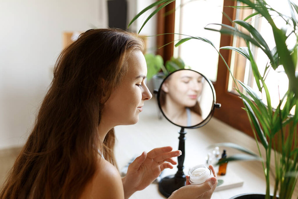 Красавица смотрит в зеркало, прикасаясь к лицу и нанося крем, морщины во время утренней процедуры красоты. Счастливая улыбающаяся красивая молодая женщина применяет увлажняющий лосьон  - Фото, изображение