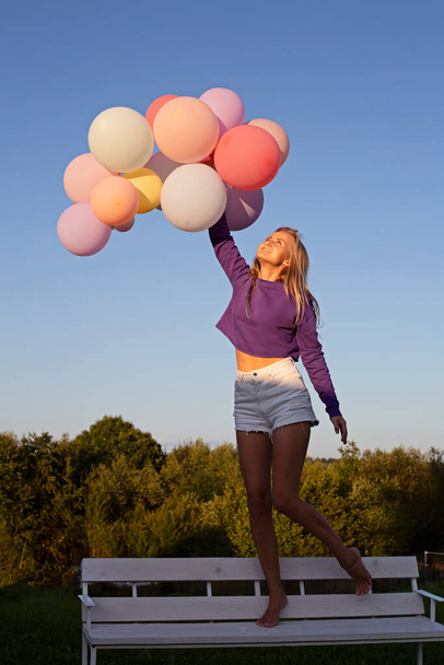 Young caucasian girl with colorful balloons on a white bench. Festive girl smiles and looks up - 写真・画像