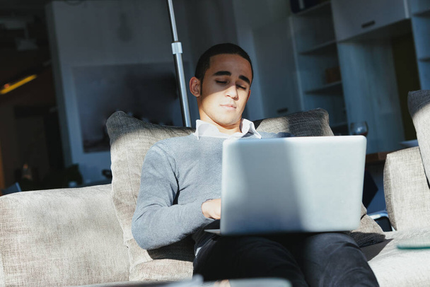 Schwarzer Mann an seinem Laptop auf einem bequemen Sofa zu Hause in einem Sonnenstrahl sitzend, während er sich auf seine Arbeit konzentriert - Foto, Bild