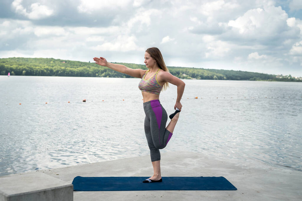 Junge schlanke Frau in Fitnessklamotten, die auf einer Yogamatte in der Nähe des Sees turnt. Konzept eines gesunden Lebensstils - Foto, Bild