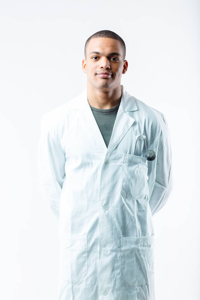 Confiado joven médico negro en bata de laboratorio blanca con estetoscopio en el bolsillo posando frente a la cámara con una sonrisa en blanco - Foto, Imagen