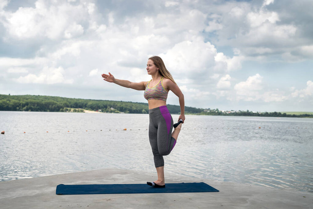 Молода біла жінка робить активні вправи на килимку для йоги біля річки влітку. Концепція здорового способу життя
 - Фото, зображення