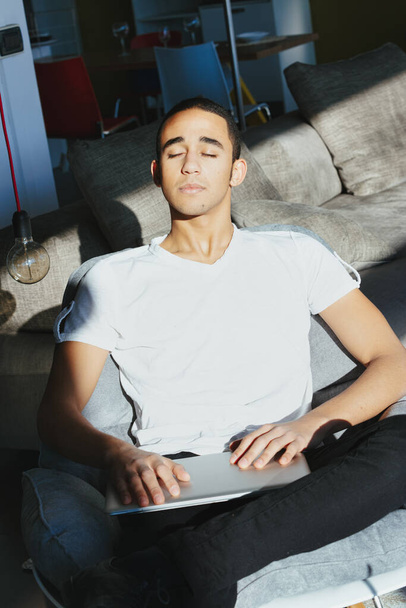 Mladý muž vychutnává teplé letní slunce, jak sedí na pohovce se zavřenýma očima a blaženým výrazem, zatímco pracuje na svém notebooku - Fotografie, Obrázek