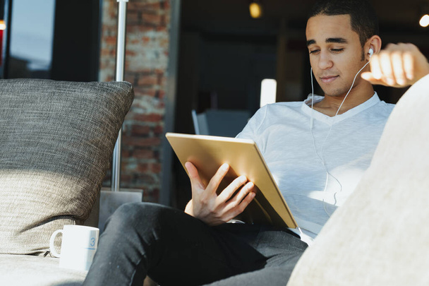 Joven hombre negro sentado escuchando música o streaming en su tablet PC con tapones para los oídos relajándose en una cómoda silla en un patio con una sonrisa contenta - Foto, imagen