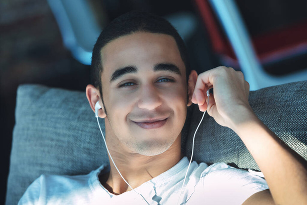 Uśmiechnięty młody, czarny mężczyzna z nieogolonym zarostem słuchający muzyki na zatyczkach do uszu w zbliżeniu, gdy relaksuje się na kanapie w słońcu - Zdjęcie, obraz