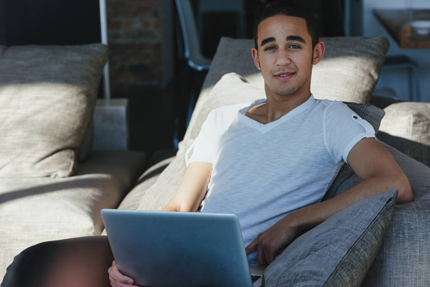Чоловік розслабляється на зручному дивані з ноутбуком у теплому сонячному світлі, дивлячись на камеру з продуманим виразом і тихою посмішкою
 - Фото, зображення