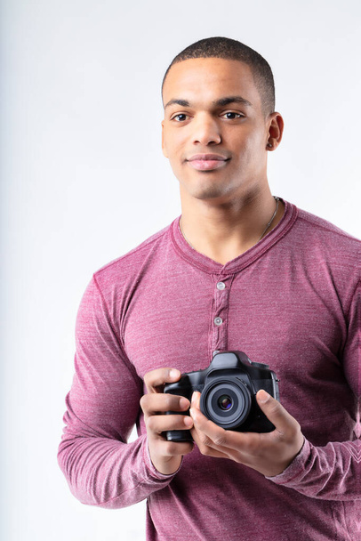 Young Black photographer standing holding his camera as he stares ahead with a quiet smile to the side of the camera in a cropped upper body view - Photo, Image
