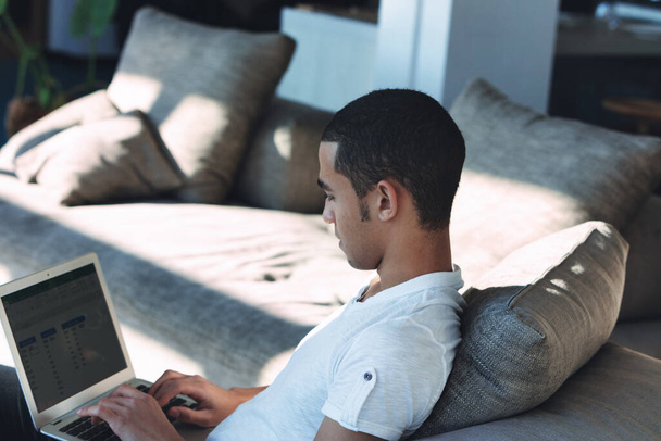 Joven hombre negro teletransportando o trabajando desde casa en una computadora portátil durante la pandemia de Coivd-19 en una vista sobre el hombro mientras se relaja en un cómodo sofá - Foto, Imagen