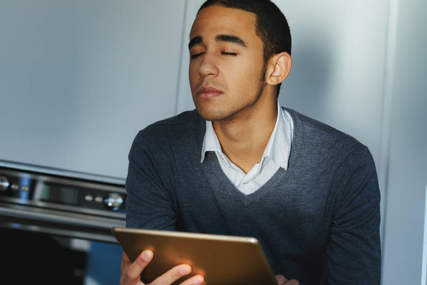 Junger Mann nimmt einen ruhigen Moment ein, um seine Gedanken zu sammeln, während er mit geschlossenen Augen und einem gelassenen Gesichtsausdruck auf einem Tablet-PC sitzt - Foto, Bild