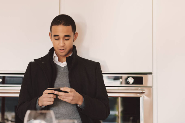 Νεαρός άνδρας με πανωφόρι στέκεται στην κουζίνα ελέγχοντας το κινητό του τηλέφωνο για μηνύματα με φόντο τους διπλάσιους φούρνους - Φωτογραφία, εικόνα