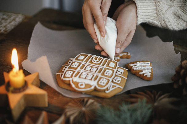 Dekorowanie świąteczne pierniki ciasteczka z lukrem na drewnianym stole ze świecą i ozdobami. Zamknij się z robieniem piernikowego domku z lukrem. Atmosferyczny obraz. Przygotowania do urlopu - Zdjęcie, obraz