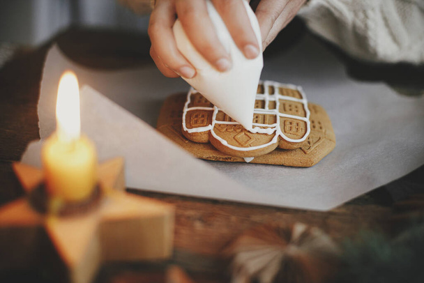 Hands decorating christmas gingerbread cookies with icing on wooden table with candle and ornaments. Close up of making gingerbread house with frosting. Atmospheric moody image. Holiday preparations - Photo, Image