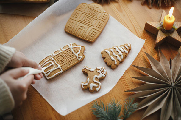 Mani che decorano i biscotti di pan di zenzero di Natale con ciliegina sul tavolo di legno con candela e ornamenti. Chiudete. Fare casa di pan di zenzero con glassa. Preparazione e tradizione delle vacanze - Foto, immagini