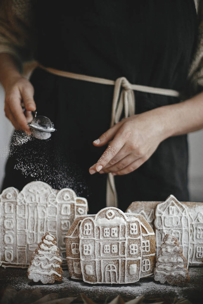 Mujer en delantal rociando azúcar en polvo en las casas de pan de jengibre de Navidad en la mesa de madera en la cocina escandinava rústica. Imagen atmosférica de mal humor. Preparación y tradiciones navideñas - Foto, Imagen