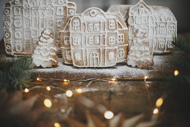 Χριστουγεννιάτικο μελόψωμο σπίτια και δέντρο σε ρουστίκ ξύλινο τραπέζι με αστέρια και φώτα. Ατμοσφαιρική κυκλοθυμική εικόνα. Χειμερινές διακοπές προετοιμασία και παραδόσεις. Καλά Χριστούγεννα! Xmas cookies χωριό σκηνή - Φωτογραφία, εικόνα