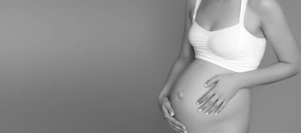 Όμορφη έγκυος νεαρή γυναίκα σε ρούχα για έγκυες γυναίκες μετρά γυμνή κοιλιά της, χαμογελώντας, σε ένα φόντο πολιορκία. Φωτογραφία μιας ευτυχισμένης εγκύου που ποζάρει πάνω από τον τοίχο. Κοιτάζοντας την κάμερα - Φωτογραφία, εικόνα