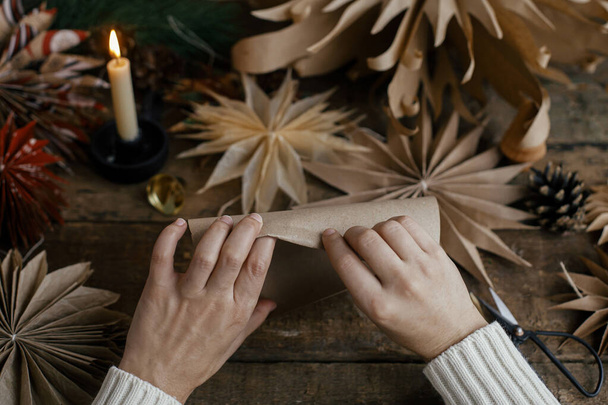 Stijlvolle kerststerren maken. Handen vouwen ambachtelijk papier op de achtergrond van handgemaakte Zweedse sterren, draad, kaars, schaar op rustiek hout. Proces van het maken van feestelijke decor. Voorbereidingen - Foto, afbeelding