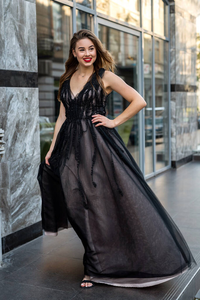 portrait of attractive female model in gorgeous evening black dress posing in street of city, summer day - Photo, Image