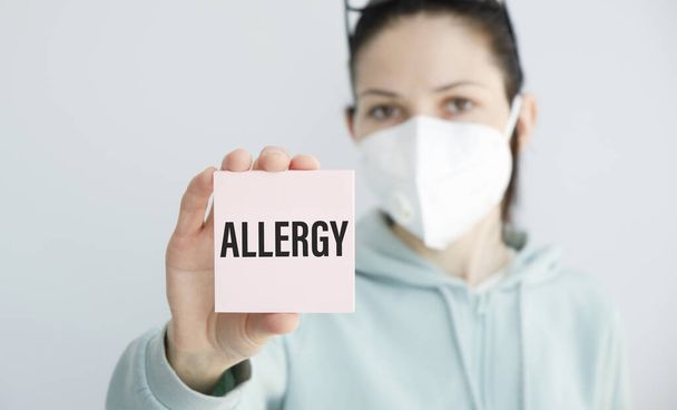 A girl in a mask holds a card in her hand with the inscription allergy - Foto, Imagen