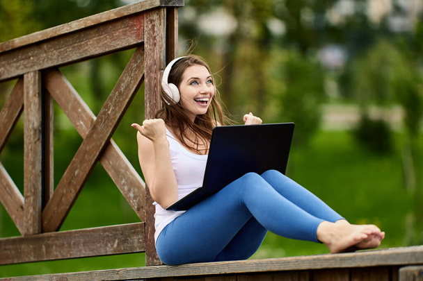 Cheerful woman in wireless headphones makes telework with laptop outdoors. - Foto, Imagen