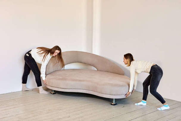 Two women in jeans move sofa in new flat. - Фото, изображение