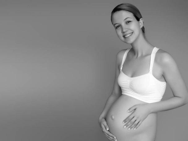 Hermosa mujer embarazada con ropa para mujeres embarazadas está midiendo su barriga desnuda, sonriendo, sobre un fondo. Imagen de la mujer embarazada feliz posando sobre la pared. Mirando la cámara - Foto, imagen