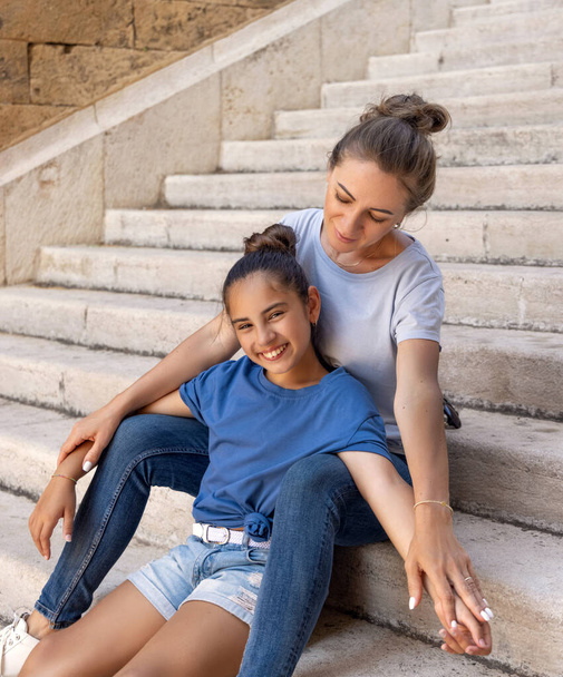 Middle-aged Mother and her kid daughter sitting on stone stairs outdoor, holding hands and looking in camera. Loving family wearing blue t-shirts and jeans spending time together outdoors in summer, t-shirts mockup - Fotó, kép