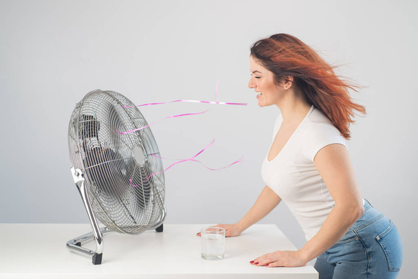 A red-haired Caucasian woman chills by the electric fan and drinks a cold drink. Climate control in the apartment - Foto, imagen