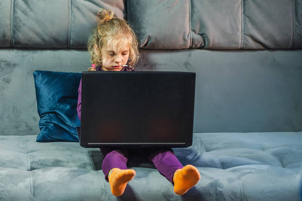 A blond-haired girl with a lollipop in her mouth sits on a gray couch with a laptop on her feet. The child's gaze is directed to the screen. The concept of a digital childhood - Foto, Imagem