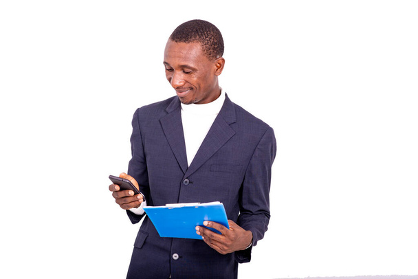 pohledný podnikatel v bundě stojí na bílém pozadí drží notepad a dívá se na mobilní telefon s úsměvem. - Fotografie, Obrázek