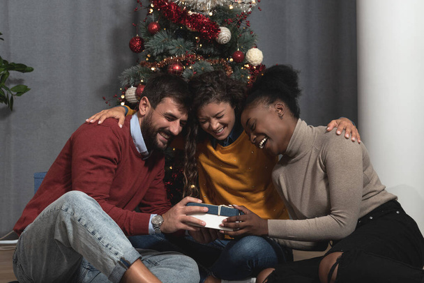 Group of young happy smiling people sitting at home with decorated Christmas tree exchanging and giving Christmas gifts celebrating Holidays and New Year Eve with warm home decoration - Zdjęcie, obraz