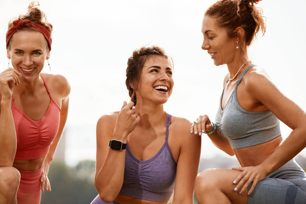 Glimlachende fitnessvrouwen in trainingskleding praten tegen elkaar op straat. Drie vrouwen joggers staan op straat tijdens hun ochtendjog - Foto, afbeelding